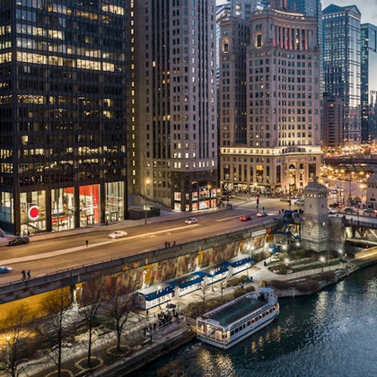 Elevated view of a cityscape at dusk showing tall buildings, a busy road, illuminated streetlights, and boats docked on a river.
