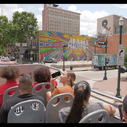 Tourists seated on an open-top bus view a colorful mural reading "Houston Inspired" in an urban street setting.