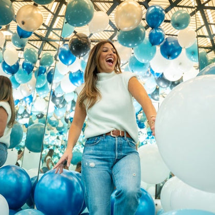 A woman smiles in a room filled with blue, white, and silver balloons, surrounded by mirrored walls and ceiling.