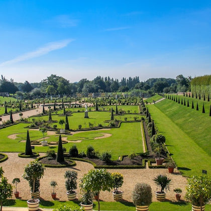 A formal garden with neatly trimmed hedges, potted plants, gravel paths, and manicured lawns under a clear blue sky.