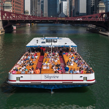 A tour boat named Skyview filled with seated passengers cruising on a river under a red bridge in a cityscape.