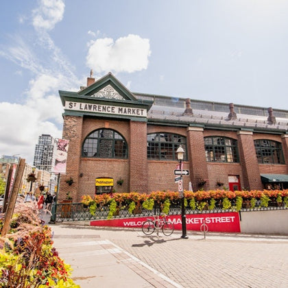 Exterior of St. Lawrence Market, a historic brick building with arched windows, surrounded by colorful flowers and signage.