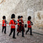 Uniformed guards, with bearskin hats and red coats, march on a cobblestone street beside a stone wall.
