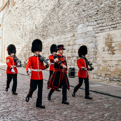Uniformed guards, with bearskin hats and red coats, march on a cobblestone street beside a stone wall.