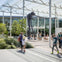 People walking in a modern outdoor plaza with greenery and a large sculpture of a figure leaning on a long stick.