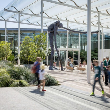 People walking in a modern outdoor plaza with greenery and a large sculpture of a figure leaning on a long stick.