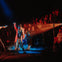 A performer in theatrical costume gestures dramatically on a smoky stage, illuminated by colorful lights, with an audience watching.
