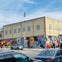 Street corner with a colorful mural covering a building, pedestrians and parked cars in view under a blue sky.