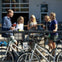 A group of people standing outdoors by a railing, holding drinks, with bicycles parked in the foreground.