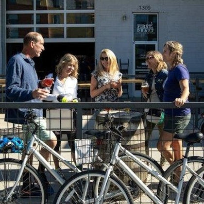 A group of people standing outdoors by a railing, holding drinks, with bicycles parked in the foreground.