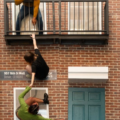 Three people appear to be climbing and hanging from a fake brick wall with a balcony, creating an illusion of danger.