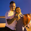 A man and woman are smiling and pointing at the camera. The woman is holding a red heart-shaped balloon.