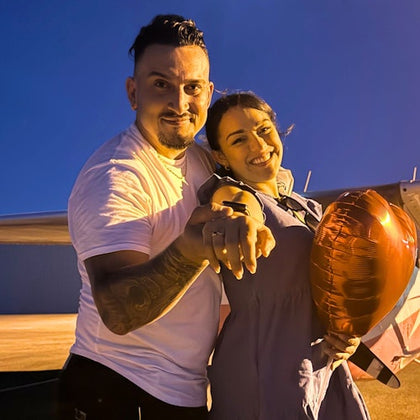 A man and woman are smiling and pointing at the camera. The woman is holding a red heart-shaped balloon.