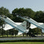 Two jet planes on display outdoors, mounted on stands, surrounded by trees and greenery on a sunny day.