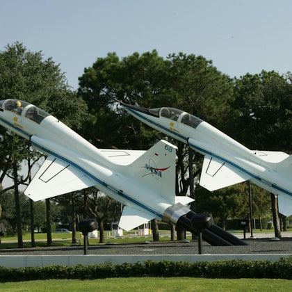 Two jet planes on display outdoors, mounted on stands, surrounded by trees and greenery on a sunny day.