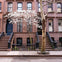 A brownstone building with two staircases, a blooming tree with white flowers, and a bare tree on the sidewalk.