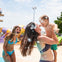 A woman holding a toddler and smiling at a water park, with a young woman in a bikini laughing nearby.