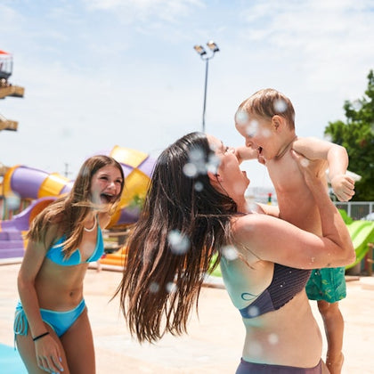 A woman holding a toddler and smiling at a water park, with a young woman in a bikini laughing nearby.