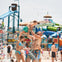 People enjoying a water park, with a man lifting a child, a woman nearby, and water slides in the background.
