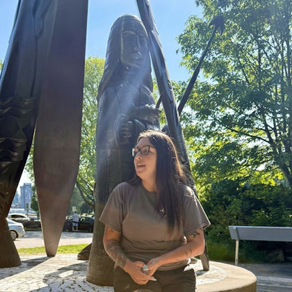 A person sits near a statue outdoors, with sunlight filtering through trees in the background.