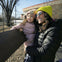 A smiling woman and child in winter clothes, including hats and jackets, stand by a fence outdoors on a sunny day.