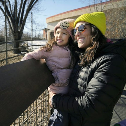 A smiling woman and child in winter clothes, including hats and jackets, stand by a fence outdoors on a sunny day.