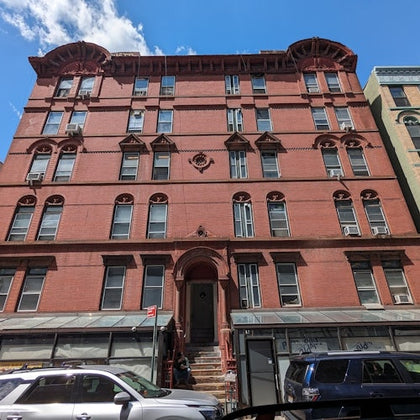 A red brick building with storefronts on the ground floor, under a blue sky with clouds. Vehicles are parked in front.