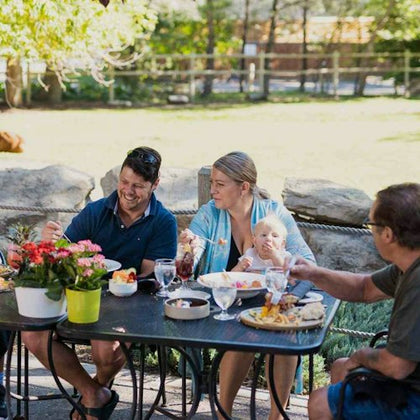 Four people, including a baby, are seated around an outdoor table enjoying a meal. The table has food, drinks, and flowers.