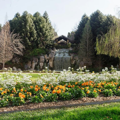 A landscaped garden with colorful flowers in the foreground, a waterfall in the center, and trees in the background.