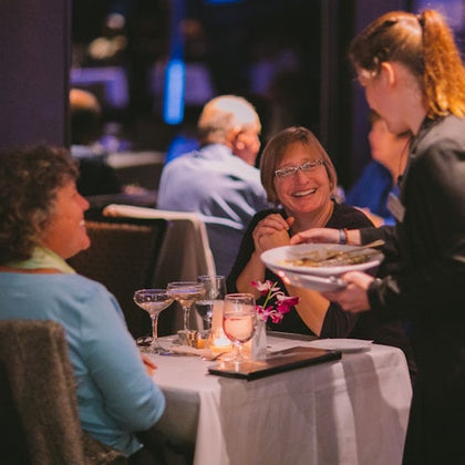 Two women seated at a restaurant table, smiling and being served by a waitress; candlelight and drinks on the table.