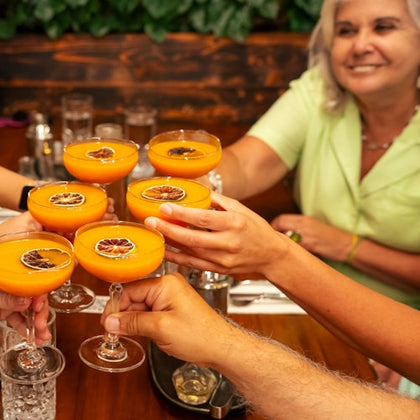 A group of people toasting with orange cocktails garnished with dried fruit, seated at a wooden table with a leafy backdrop.