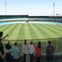 A group of people standing in a stadium, viewing a large green field with goal posts under a clear sky.