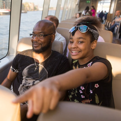 A man and a girl sitting on a boat, with the girl pointing forward and smiling. The background shows water and other passengers.