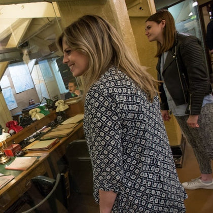 Two women observe an old-fashioned office setup through a glass partition, featuring a desk with vintage items.