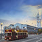 A double-decker tour bus drives past a clock tower and waterfront building under a partly cloudy sky.