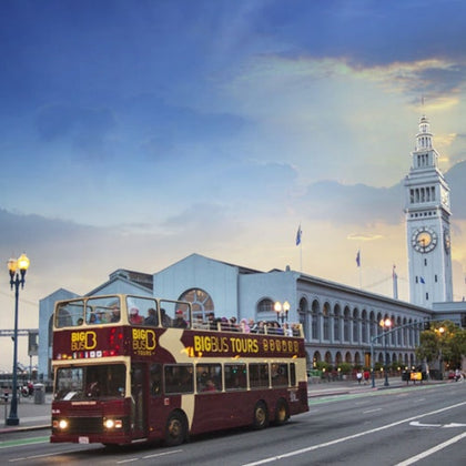 A double-decker tour bus drives past a clock tower and waterfront building under a partly cloudy sky.