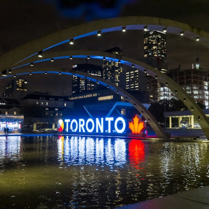 The Toronto sign at Nathan Philips Square lit up at night and framed by the concrete arches above the water feature