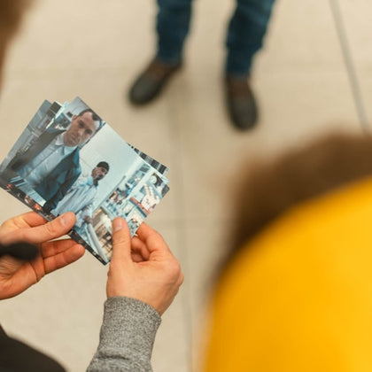 Hands holding a photo of two men, man in foreground in formal attire. Floor tiles and legs of another person visible in the background.