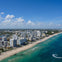 Aerial view of a coastline with a sandy beach, turquoise ocean, and a cityscape featuring tall buildings under a blue sky.