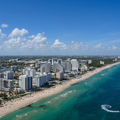 Aerial view of a coastline with a sandy beach, turquoise ocean, and a cityscape featuring tall buildings under a blue sky.