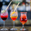 Three glasses of colorful cocktails with straws on a bar surface, with a blurred background of bottles and lights.