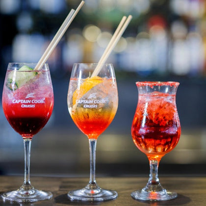 Three glasses of colorful cocktails with straws on a bar surface, with a blurred background of bottles and lights.