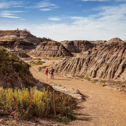 Two people walking on a dirt path amidst rugged, dry rock formations under a blue sky with light clouds.