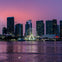 City skyline at sunset with tall buildings, a Ferris wheel, and reflections on the water in the foreground.