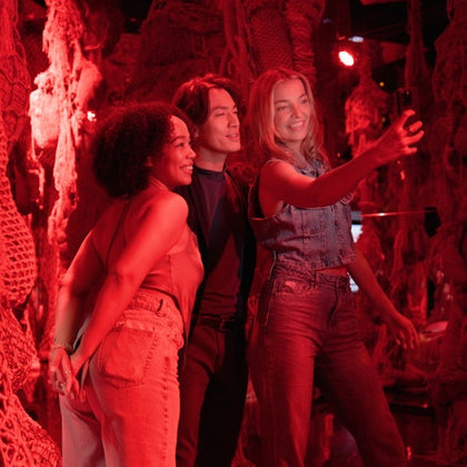 Three friends take a selfie in a dimly lit, red-hued room filled with hanging net decorations.