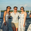 Three people smiling and posing together on a boat deck with a body of water and sailboats in the background.