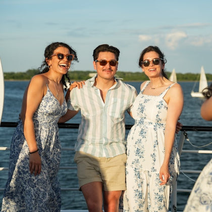 Three people smiling and posing together on a boat deck with a body of water and sailboats in the background.