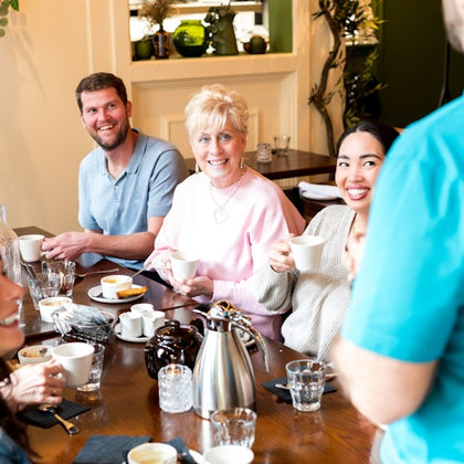 Three people are sitting at a dining table, smiling and holding coffee cups. The table has drinks and pastries on it.