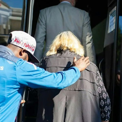 A person in a blue jacket and cap assists a blonde-haired individual wearing a black vest as they board a bus.