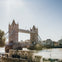 Tower Bridge of London spanning the River Thames on a sunny day, with people walking along the riverside.